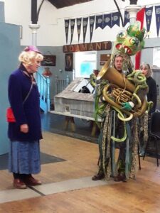 Photo of 2 people rehearsing for a performance of Rapunzel--Lockdown, by Cyclops Productions, London 2024. Left, Andrew Hodgson, dressed as a woman, playing the part of Dame Gothel. On the right, Dave Powell, in the role of Kraken, playing a tuba and wearing green tatters, a very large sea monster head with tentacles and great big goggly eyes. Photo S. Kingshill