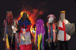 5 people in colourful Guiser folk play costumes on a dark night with a bonfire behind them; they are dressed as typical hero play characters, including a knight with a St George's cross tabard and a wooden sword; a doctor in a top hat; a woman with a basket; each person's face is completely covered with scarves or chain mail, etc.