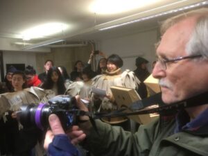 man with a moustache & glasses & a large camera filming young people mummer's costumes of tattered brown paper