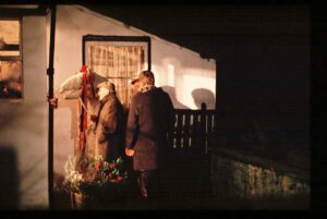 A Mari Lwyd: a person under a sheet holds a beribboned horse skull on a stick, beside 3 people in winter clothes standing outside a house at night waiting for the door to be opened 