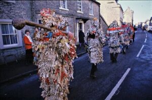 rear view of about 7 people walking up the middle of a road wearing tattered costumes made of coloured shreds of paper