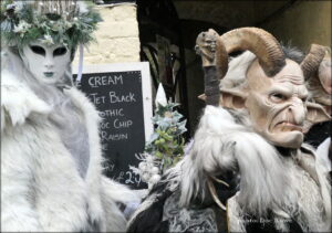 a person on the left wearing a white fur costume, a white canrival mask, and a head dress of greenery and white flower; on the right, a person in a mask with a long nose and pointed ears, a head dress of ram's horns, and a fur-collared jacket