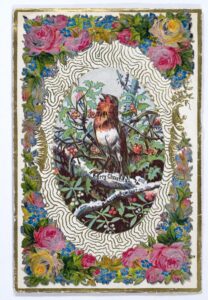 Victorian Christmas card in The Folklore Society Archives, showing a singing robin on a branch surrounded by greenery, in a white oval with a lace effect, and bordered with pink flowers and greenery 