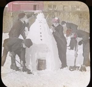 Hand-coloured photograph magic lantern slide from around the 1910s showing a group of 6 boys making a large snowman, with shovels and hands. The snowman has coals or stones for face and buttons, and at its base is a school slate with the chalked message 'Glad to See You.' Digital photo Nigel Longstaff. 