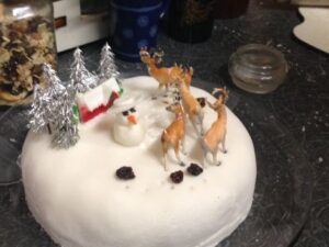 Photo of a Christmas cake with royal icing, decorated with: 5 silver Christmas trees, a small model of a snow-covered cottage, a snowman bigger than the house, 4 plastic reindeer, reindeer hoofprints in the icing and 4 lumps of brown cake representing reindeer droppings